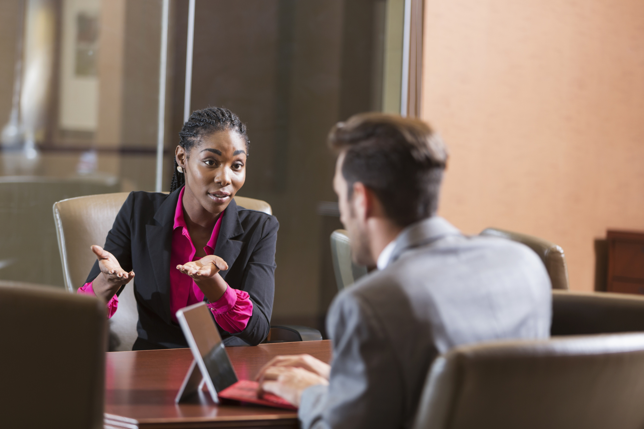 A young, serious African American woman sitting in a boardroom across the table from a young businessman. They are both wearing business suits and the man is looking at the woman and typing on a digital tablet. She is in focus and may be answering questions for a job interview.