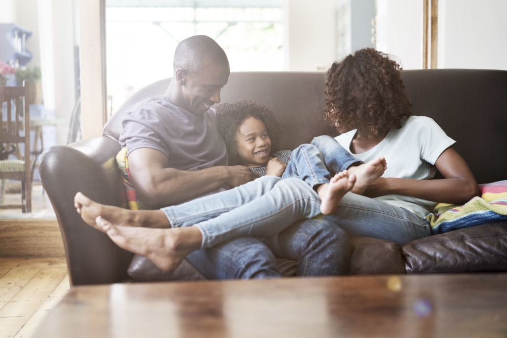A photo of cheerful family sitting on sofa at brightly lit home. Happy parents are playing with son in living room. They are in casuals.